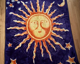 May include: A rectangular plush throw blanket featuring a deep purple background with a large central sun graphic. The sun has a human-like face in yellow and orange with radiating wavy rays. The design includes scattered orange stars and a crescent moon.