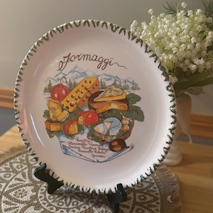 Puede incluir: Un plato de cerámica blanco con un borde festoneado verde y una ilustración de queso. El plato presenta la palabra "Formaggi" en cursiva y una variedad de quesos, incluyendo tomates rojos y un paisaje montañoso.