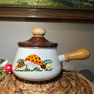 Könnte beinhalten: Ein weiß gesprenkelter Emaille-Topf im Vintage-Stil mit braunem Deckel und Holzgriff. Der Topf hat ein skurriles Pilzdesign in Orange, Gelb und Braun. Der Topf steht auf einem gewebten Platzset.
