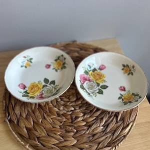 Könnte beinhalten: Zwei cremefarbene Keramikplatten mit einem Rosendesign in Gelb, Rosa und Weiß mit grünen Blättern. Die Teller haben einen goldenen Rand. Sie liegen auf einem gewebten, braunen Platzset.
