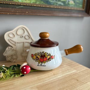 May include: White enamel fondue pot with a brown lid and wooden handle. The pot features a floral design with red tomatoes, mushrooms, and green leaves. The text "La Fondue" is printed on the front of the pot.