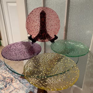Può includere: Quattro piatti di vetro colorati con una superficie strutturata a forma di bolle. I piatti sono nelle tonalità del rosa, del viola, del verde e del giallo. I piatti sono esposti su un tavolo di vetro con una base in legno.