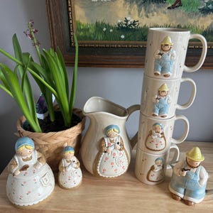 May include: A vintage set of ceramic kitchenware featuring a family of figurines. The set includes a jug, two salt and pepper shakers, and four mugs. The figurines are all dressed in simple clothing and have a whimsical, folk art style. The mugs are stacked on top of each other, and the jug is in the centre of the image.