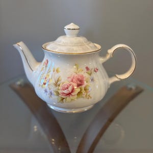 以下が含まれることがあります： 蓋、注ぎ口、取っ手が付いた白い磁器のティーポット。 ティーポットはピンクのバラと金のトリムで装飾されています。 ティーポットはガラスの表面に置かれています。