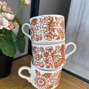 May include: Three white ceramic coffee mugs with a brown floral pattern. The mugs are stacked on top of each other.