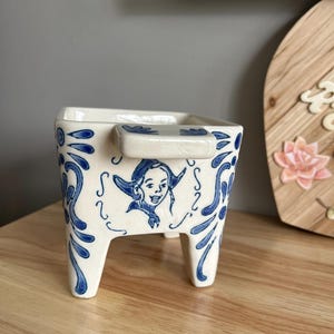 May include: A white ceramic planter with blue floral and figure designs. The planter has a square shape, three legs, and a handle. The blue designs include a person in a hat and decorative swirls. The planter is on a wooden surface.