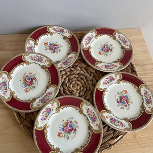 May include: Five antique porcelain plates. Each plate has a burgundy rim with a gold filigree design and a central floral bouquet. The plates are arranged on a woven mat, showcasing their intricate details and vintage charm.