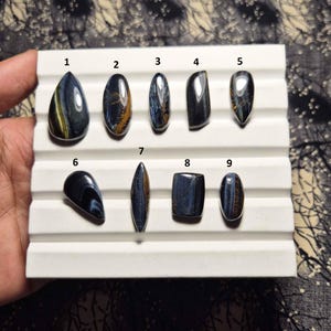 May include: Nine polished tiger eye gemstones in various shapes and sizes are displayed on a white tray. The gemstones feature a mix of blue, gold, and brown colors, with unique patterns and striations. The gemstones are numbered 1 through 9.