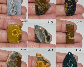 May include: Nine different gemstone cabochons, each with unique patterns and colours, including blue, green, black, brown, and yellow. The cabochons are displayed on a hand.