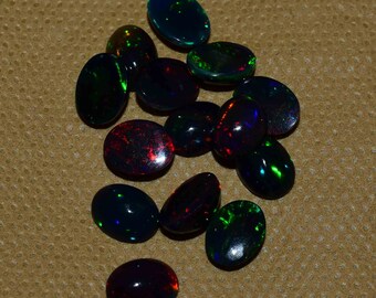 May include: A collection of 14 oval-shaped black opals with iridescent green, blue, and red flashes. The opals are arranged in a loose pile on a brown textured surface.