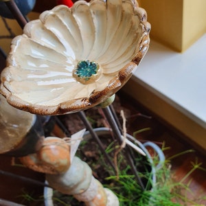 以下が含まれることがあります： 茶色の縁取りと緑色の中心を持つ、花の形をしたセラミック製のバードバス。バードバスは金属製のスタンドに取り付けられており、緑色の植物に囲まれています。