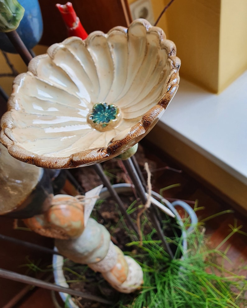 以下が含まれることがあります： 茶色の縁取りと緑色の中心を持つ、花の形をしたセラミック製のバードバス。バードバスは金属製のスタンドに取り付けられており、緑色の植物に囲まれています。