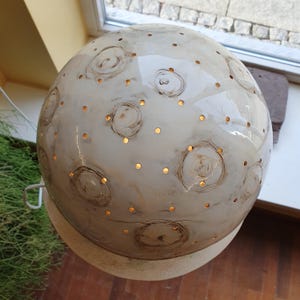 Może przedstawiać: Kulista, kremowa lampa ceramiczna z błyszczącym wykończeniem. Lampa posiada wzory spiralne i kropkowe, z małymi otworami emitującymi ciepłe, złote światło. Lampa stoi na okrągłej, kremowej podstawie.