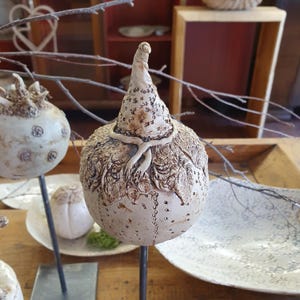 May include: A collection of handcrafted ceramic sculptures. The central piece is a beige, textured sculpture with a conical hat and a decorative scarf. Other sculptures include a round, textured piece and a small, white, round sculpture. The sculptures are on metal stands.