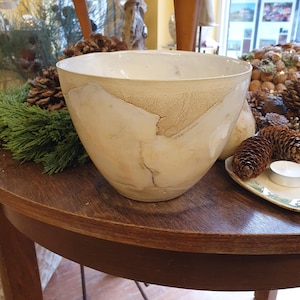 May include: A large, off-white ceramic bowl with a wide opening. The bowl has a textured, beige band around the top and a crack pattern. The bowl sits on a dark wooden table, with pine cones and greenery in the background.