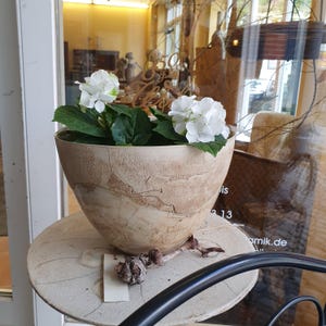 May include: A decorative, light-colored, ceramic bowl with a textured surface, filled with green leaves and white hydrangea flowers. The bowl sits on a round, light-colored, ceramic stand. A small piece of wood and a tag are on the stand.