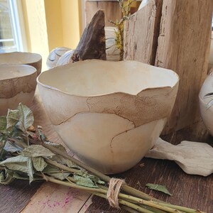 May include: A ceramic bowl with an off-white glazed interior and a textured, matte tan exterior rim. The bowl features an irregular, organic shape with a wavy top edge. The surface displays a contrast between the smooth, glossy finish and the rough, earthy clay texture along the upper portion of the vessel.