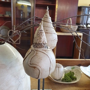 May include: Two off-white ceramic sculptures with textured surfaces and unique shapes. One is pear-shaped with a spiral design, and the other has a bulbous base and a textured top. They are displayed with branches and other decorative items.