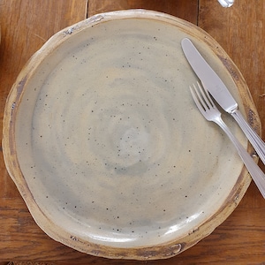 Könnte beinhalten: Ein großer, runder Teller aus Keramik mit einem gesprenkelten, hellgrauen Überzug. Der Teller hat einen leicht erhöhten Rand und ein subtiles, wirbelndes Muster. Zwei silberne Besteckteile liegen auf dem Teller.