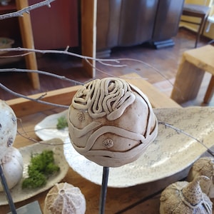 May include: A beige ceramic sculpture with a textured, brain-like design, mounted on a dark metal rod. The sculpture has raised, swirling patterns and small, circular details. Other ceramic pieces are visible in the background, suggesting a collection of decorative art.