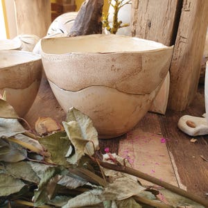 May include: A hand-formed ceramic bowl with an irregular, wavy rim. The bowl features a two-tone finish with a smooth, cream-colored lower section and a textured, speckled brown upper band that follows a curved line around the exterior surface.