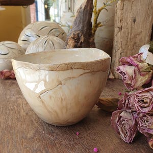 May include: A hand-formed ceramic bowl with an irregular, wavy rim. The bowl features a glossy, off-white glaze with visible brown, marbled streaks and a textured, earthy brown band along the upper edge, showcasing a unique, organic shape.