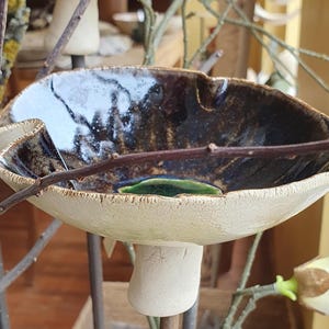 May include: A ceramic mushroom-shaped sculpture featuring a wide, off-white textured cap with a glossy, dark brown interior glaze and a small, vibrant green accent at the centre. The stem is a simple, matte off-white cylinder supporting the cap.