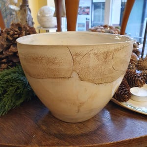 May include: A large, off-white ceramic bowl with a textured, brown band around the upper half. The bowl has a wide opening and a rounded base. The surface has visible cracks, adding to its rustic charm. The bowl is likely handmade.