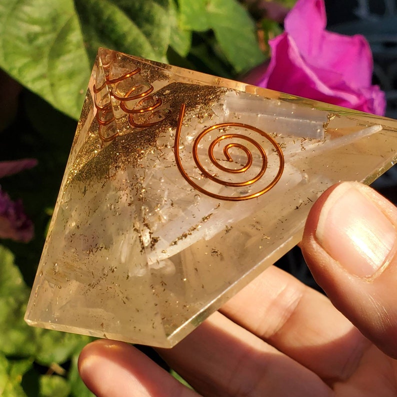 May include: A clear quartz crystal pyramid with a copper wire spiral design. The pyramid is sitting on a hand.