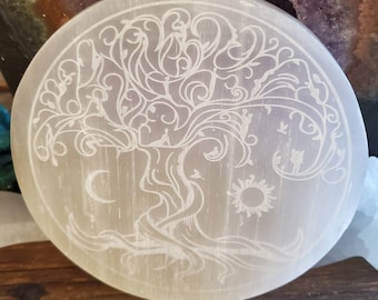 May include: A white selenite charging plate with a tree of life design etched into the surface. The tree has intricate branches and leaves, and there is a crescent moon and sun at the base of the trunk.