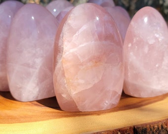 May include: Several polished rose quartz stones, each in an egg-like form. The stones exhibit a delicate pink colour with faint internal patterns. They are presented on a natural wooden base with a bark edge, creating a natural aesthetic.