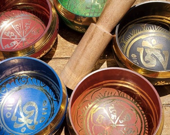 May include: Set of five singing bowls with a wooden striker. The bowls are made of metal and have intricate designs and lettering in gold. The bowls are in various colours, including red, blue, green, and black.