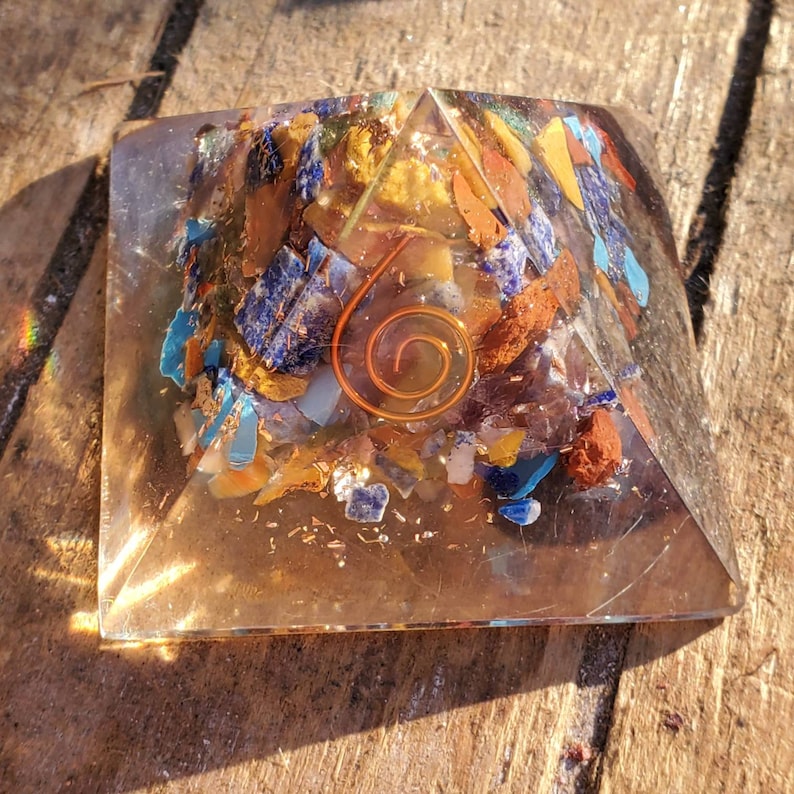 K&ouml;nnte beinhalten: Eine klare Kristallpyramide mit einer Kupferspirale im Inneren. Die Pyramide ist gef&uuml;llt mit kleinen St&uuml;cken verschiedener farbigener Edelsteine, darunter Blau, Gelb, Orange und Rot.