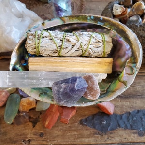 May include: A white sage smudge stick and palo santo stick are placed in a large abalone shell with a selenite wand and various crystals.