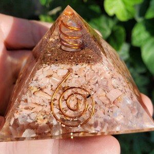 K&ouml;nnte beinhalten: Eine klare Quarzpyramide mit einer eingebetteten Kupferspirale. Die Pyramide ist mit kleinen Rosenquarzst&uuml;cken und Kupferflocken gef&uuml;llt.