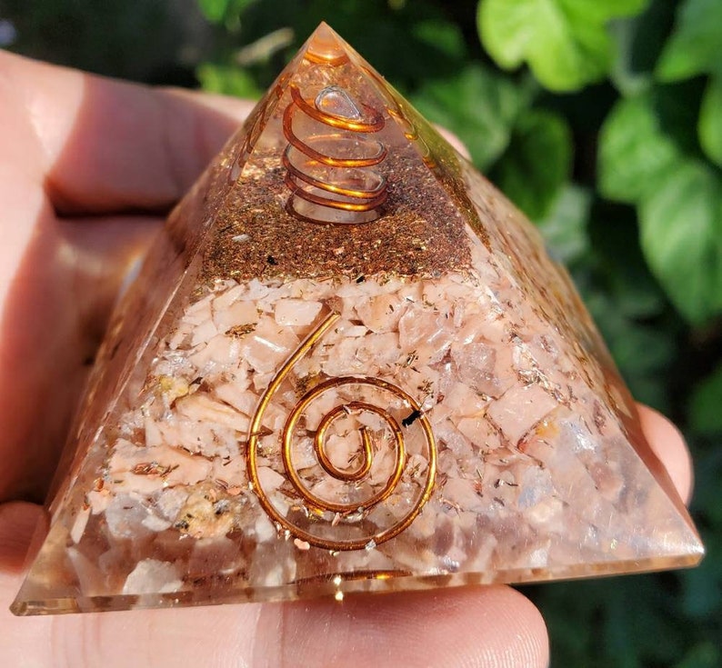 K&ouml;nnte beinhalten: Eine klare Quarzpyramide mit einer eingebetteten Kupferspirale. Die Pyramide ist mit kleinen Rosenquarzst&uuml;cken und Kupferflocken gef&uuml;llt.