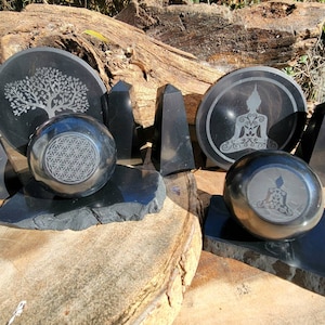 May include: Black obsidian coasters with various engraved designs, including a tree of life, a flower of life, and a meditating Buddha. The coasters are displayed on a wooden surface.