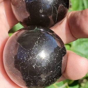 May include: Two black obsidian spheres, polished to a high shine, with a subtle sparkle. The spheres are held in a hand, showcasing their smooth, round shape.