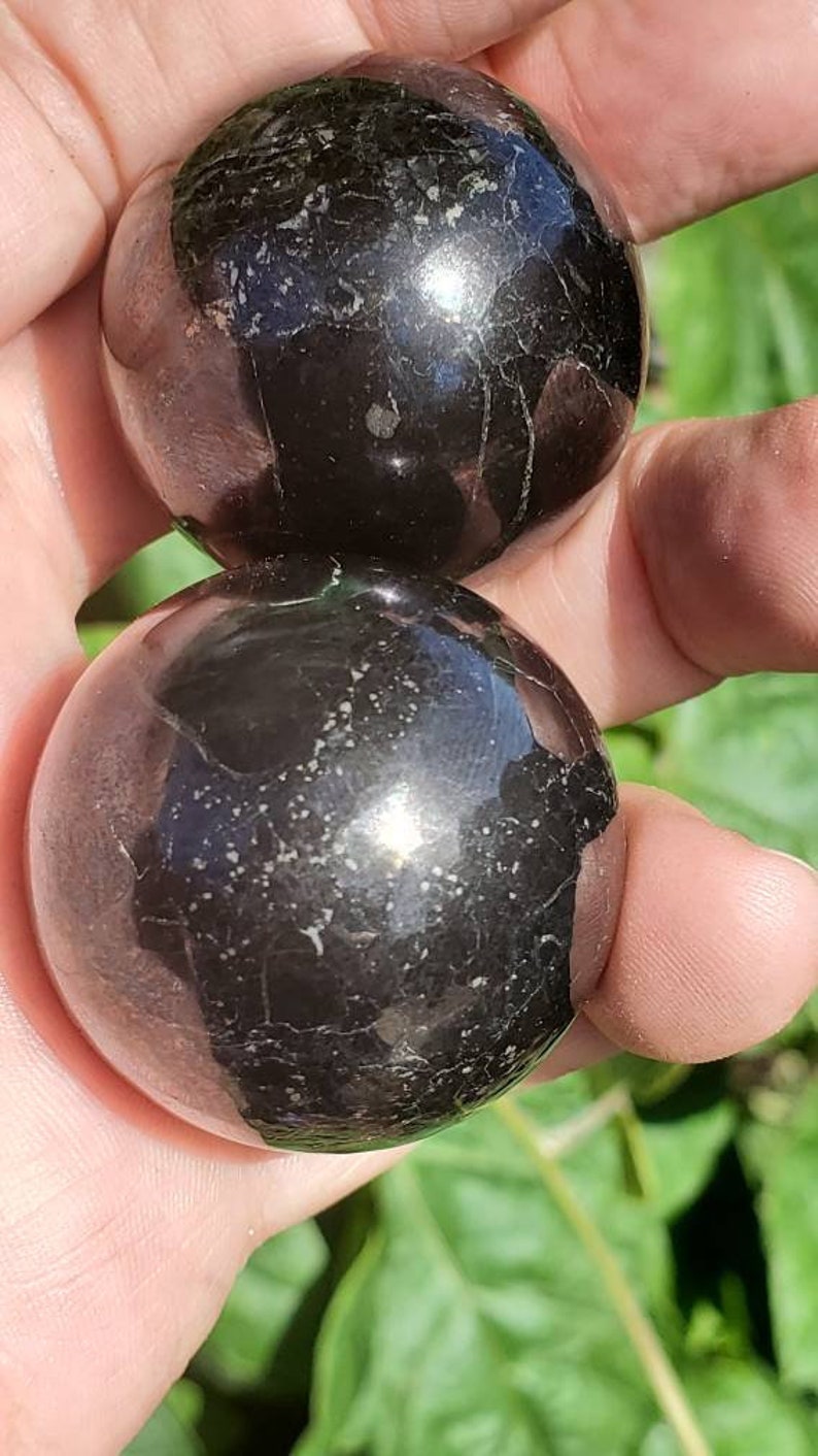 May include: Two black obsidian spheres, polished to a high shine, with a subtle sparkle. The spheres are held in a hand, showcasing their smooth, round shape.
