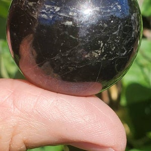 May include: A black obsidian sphere with a smooth, polished surface. The sphere has a slight sheen and is held in a hand.
