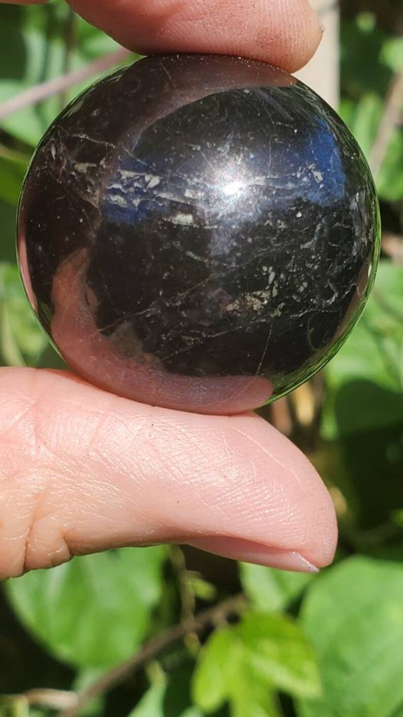 May include: A black obsidian sphere with a smooth, polished surface. The sphere has a slight sheen and is held in a hand.