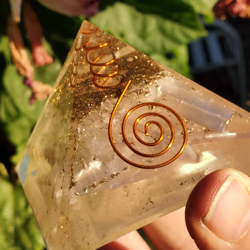 May include: A clear quartz crystal pyramid with a copper wire spiral design. The pyramid is filled with gold glitter and has a light pink hue.
