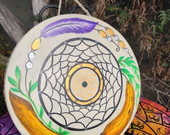 May include: A hand-painted drum featuring a dreamcatcher design. The drum is white with black and gold accents. The dreamcatcher has a web of black lines with a white centre and a gold circle around it. There are two yellow feathers and a purple feather on the drum.