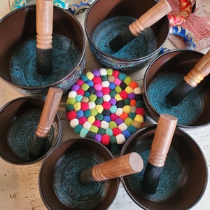 May include: Seven singing bowls with wooden strikers, arranged in a circle on a white surface. The bowls are black with a teal blue interior and intricate designs. A colorful felt circle is in the center of the arrangement.