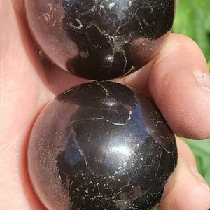 May include: Two black obsidian spheres, each with a smooth, polished surface and intricate patterns of natural inclusions. The spheres are held in a hand, showcasing their size and detail.