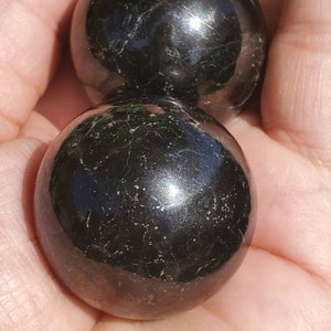 May include: Two black obsidian spheres, polished to a high shine, with a slight sparkle. The spheres are smooth and round, and they are held in a hand.