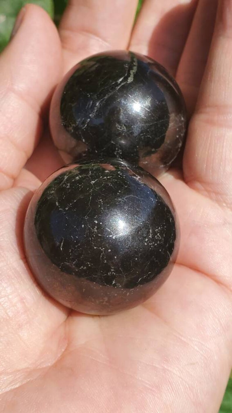 May include: Two black obsidian spheres, polished to a high shine, with a slight sparkle. The spheres are smooth and round, and they are held in a hand.
