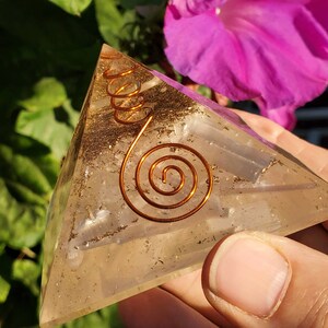 May include: Clear quartz crystal pyramid with a copper wire spiral design. The pyramid is sitting on a hand.