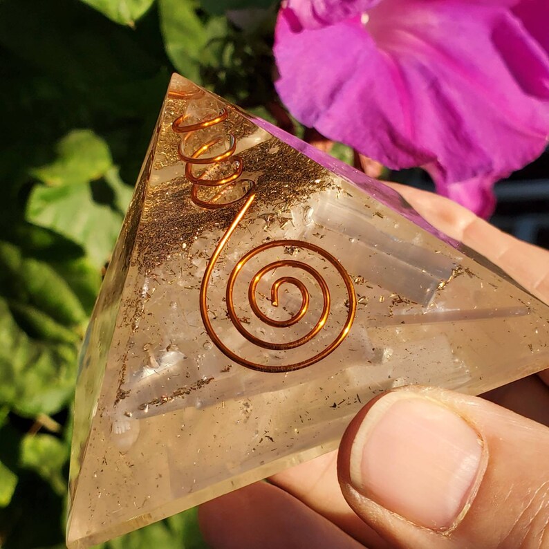 May include: Clear quartz crystal pyramid with a copper wire spiral design. The pyramid is sitting on a hand.