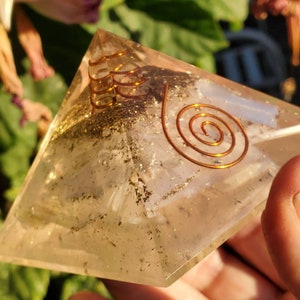 May include: A clear quartz pyramid with a copper wire spiral design. The pyramid is filled with small, sparkly particles.
